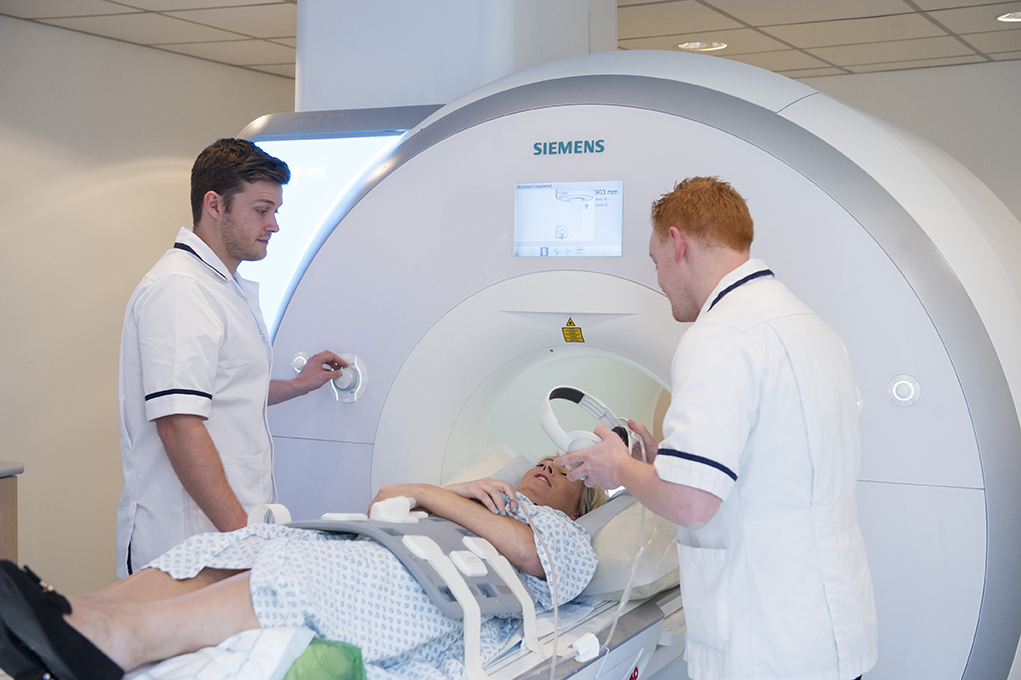 Images shows a female patient lying on the MRI bed. Two male radiographers are helping her and giving her the headphones to wear during the scan.