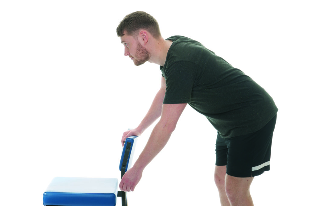 Photograph of a person leaning against a chair with one arm on the chair and one arm swung forward