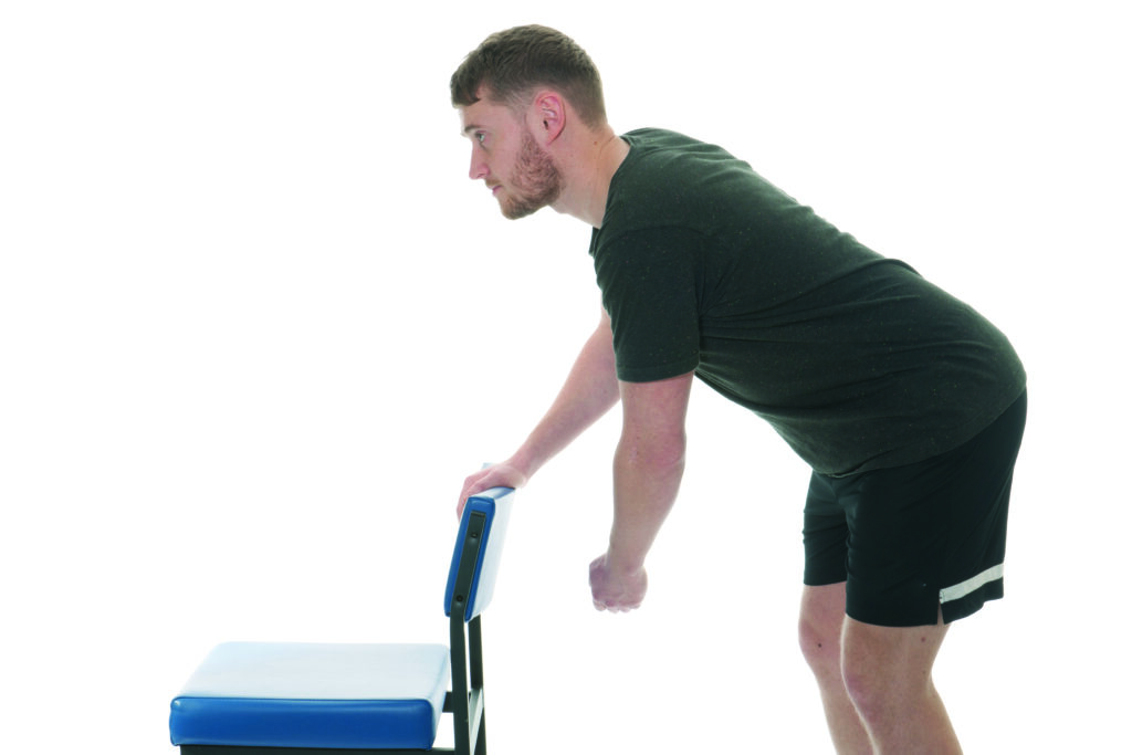 Photograph of a person leaning against a chair with one arm on the chair and one arm swung to the side