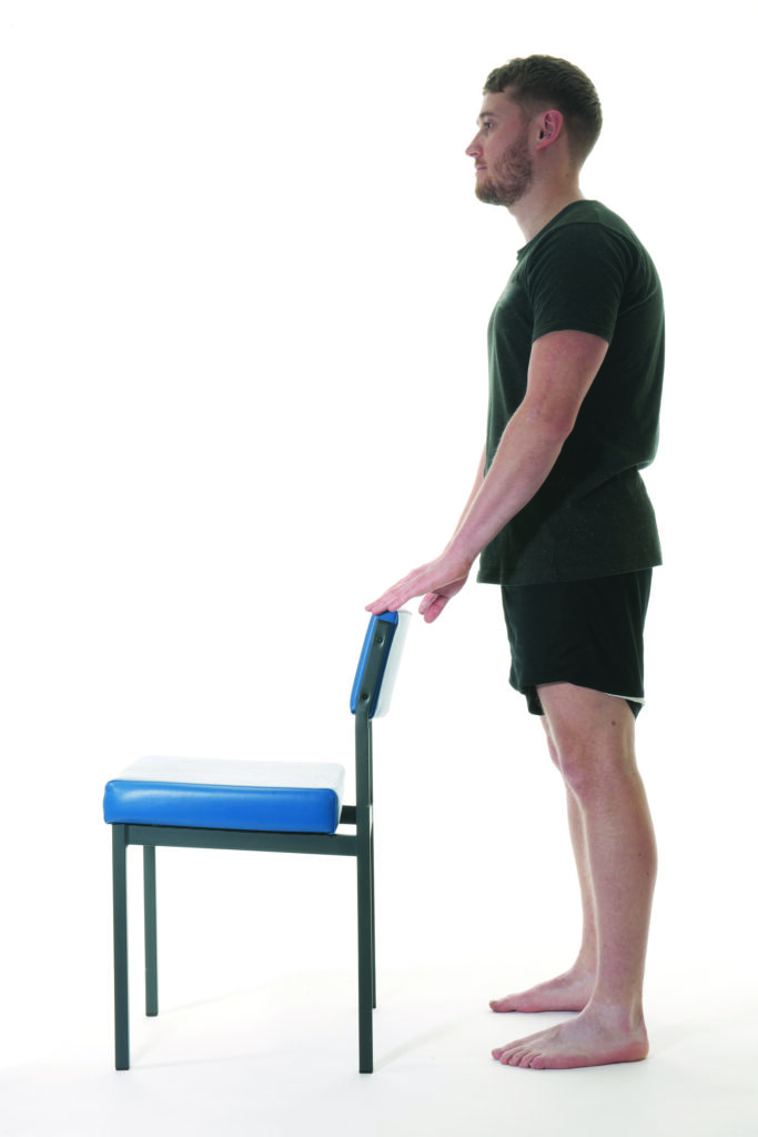Photograph of a person stood upright holding the back of a chair