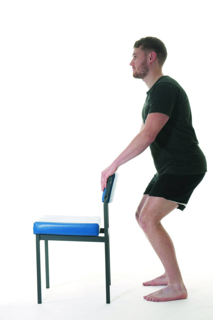Photograph of a person stood behind a chair squatting with one leg raised using the chair for support