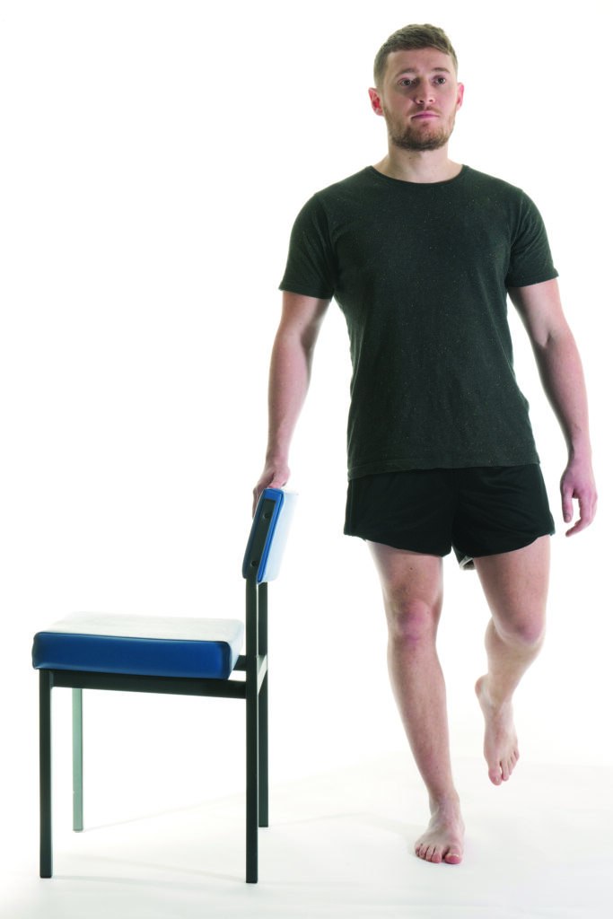 Photograph of a person stood behind a chair side on relaxed with one leg raised