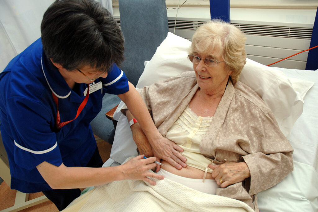 Photograph of a patient in bed with a nurse giving an injection