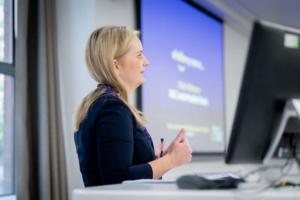 A photo of Esther Wakeman presenting at the podium with a slide with her name and title on the screen.