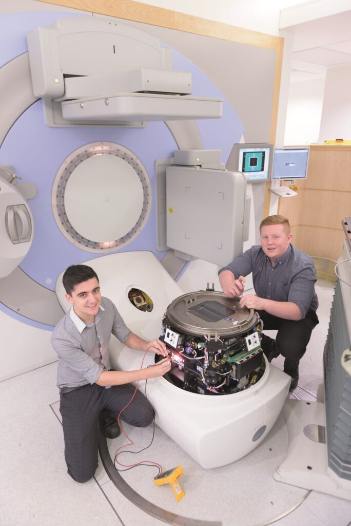 An image showing 2 males kneeling next to healthcare equipment who are fixing it. They are undertaking the Healthcare Scientist Apprenticeship at the Leeds Teaching Hospitals NHS Trust, which is run through the Medical Engineering Department.