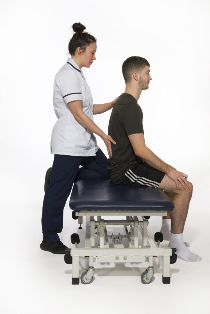 Photograph of a person sat on a bed with a physiotherapist supporting them.