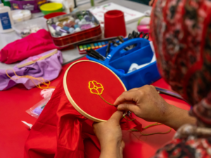 Photo at a Maternal Journal class showing a female in a red patterned hijab sewing a yellow flower on to a circular wooden frame of red fabric.