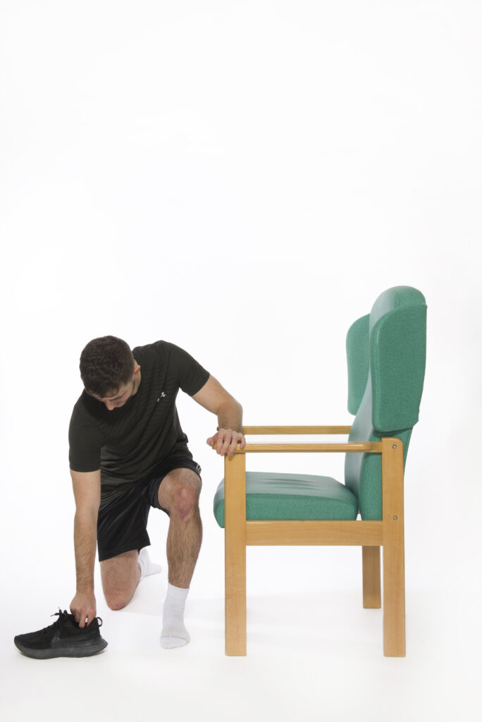 Photograph of a person bending down using a chair for support to pick up a shoe.