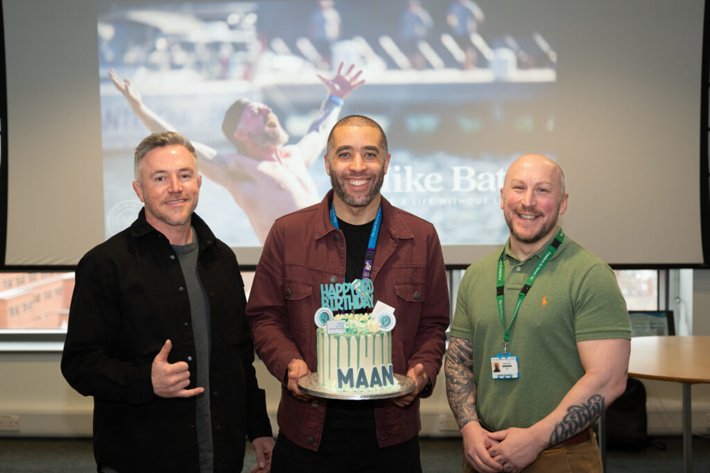 Three men stood in front of a projector screen holding a birthday cake.