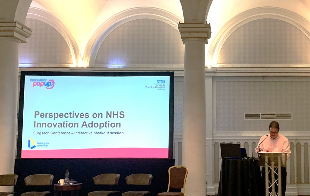 Dr Richard Evans presenting at the SurgTech conference with a screen with a slide on that reads "Perspectives on NHS Innovation Adoption"