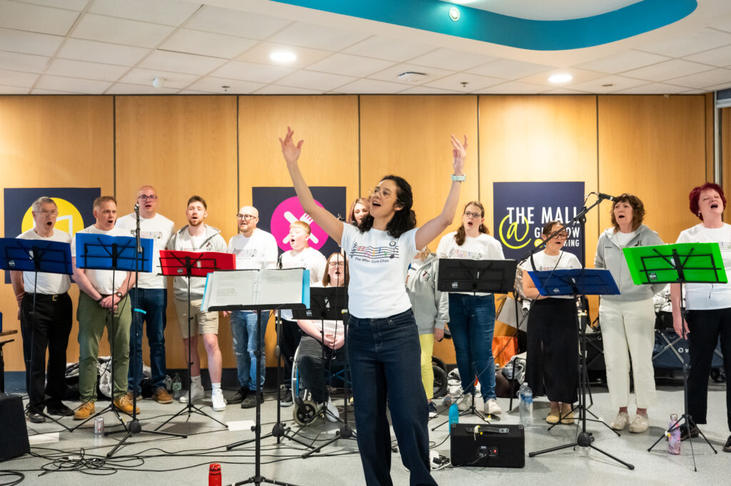 A choir stood singing from song sheets with the conductor stood arms in air facing the crowd