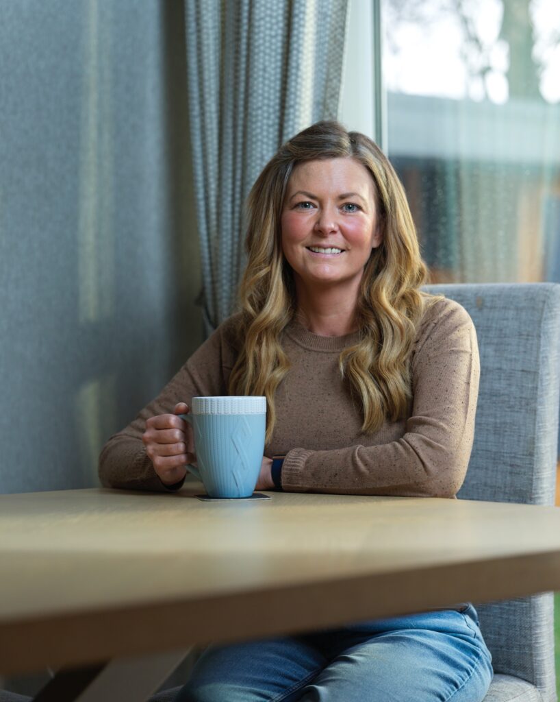 A woman sat  on a grey chair at a wooden table with a blue mug