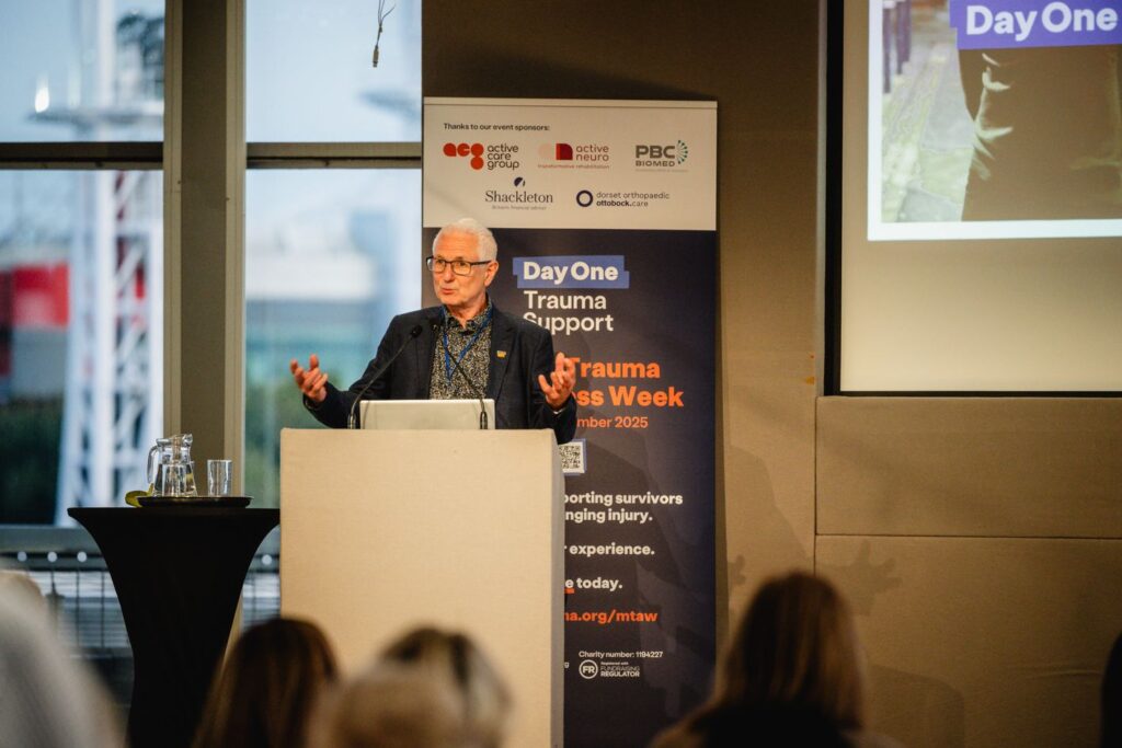 A man in a black suit jacket speaking behind a white podium in front of a room of people. Behind him is a banner for Day One Trauma Support's Major Trauma Awareness Week