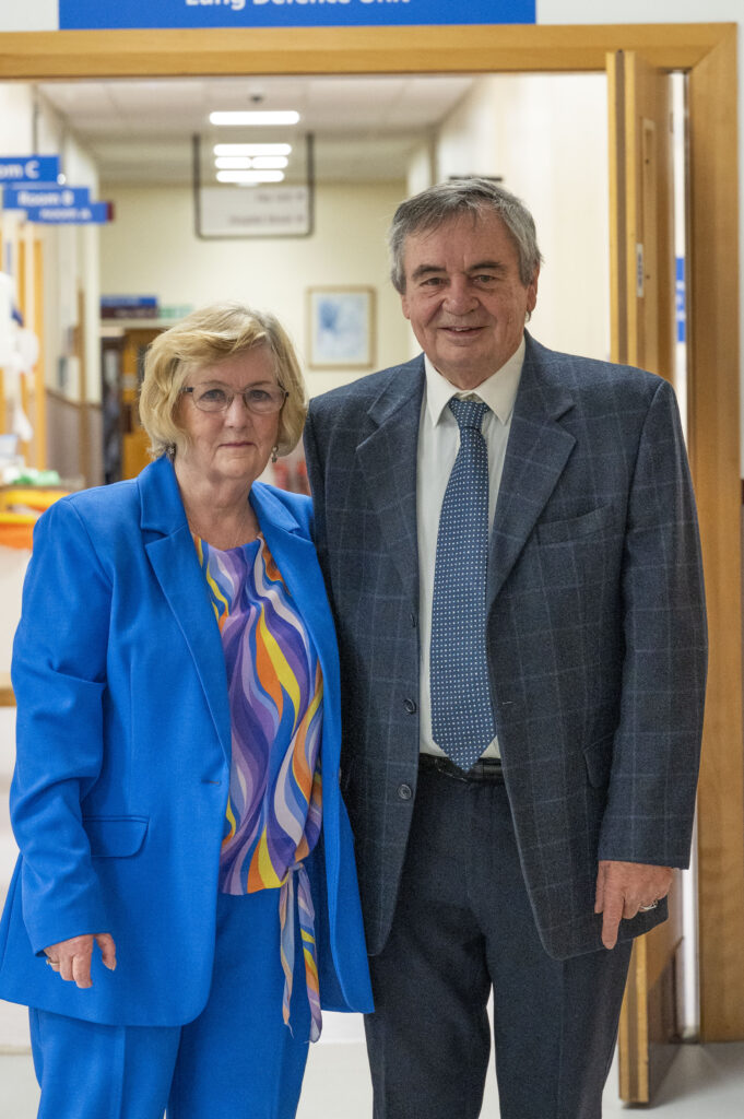 A man in a grey suit and blue tie stood with a woman in a blue suit and blouse stood in a hospital corridor