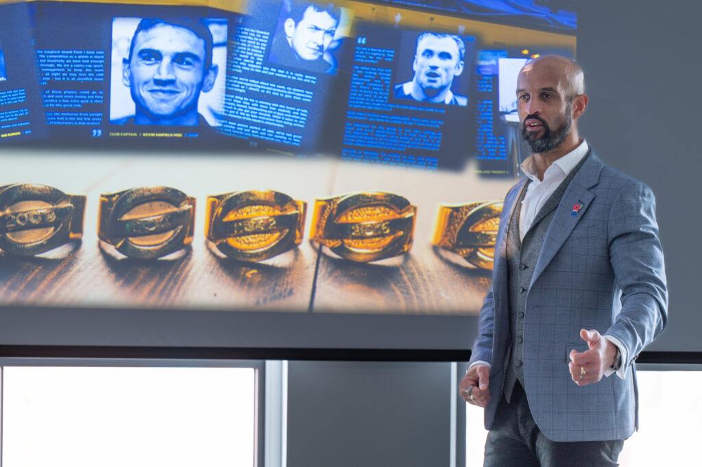 A man in a grey suit speaking in front of a display screen with pictures of gold rings