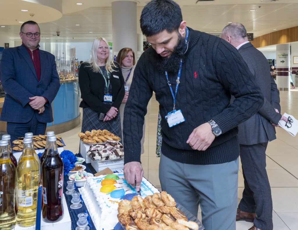 A man in cutting an iced cake on a table with pastries, drinks and other treats while others look on.