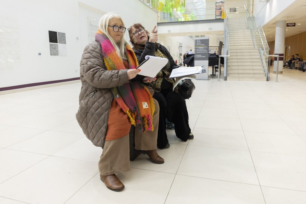 Two women in coats and scarves sat on a bench with clipboards in a hospital lobby.