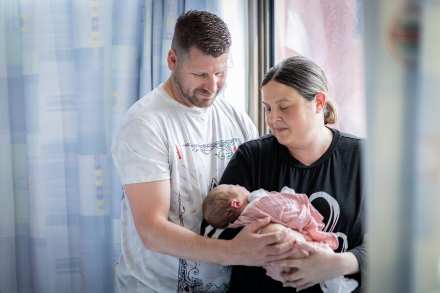 Mum in a black t-shirt stood next to a window in hospital holding a newborn baby in a pink babygrow. Dad is stood next to her in a white t-shirt - he also has his hand around the baby and they are both looking at her.