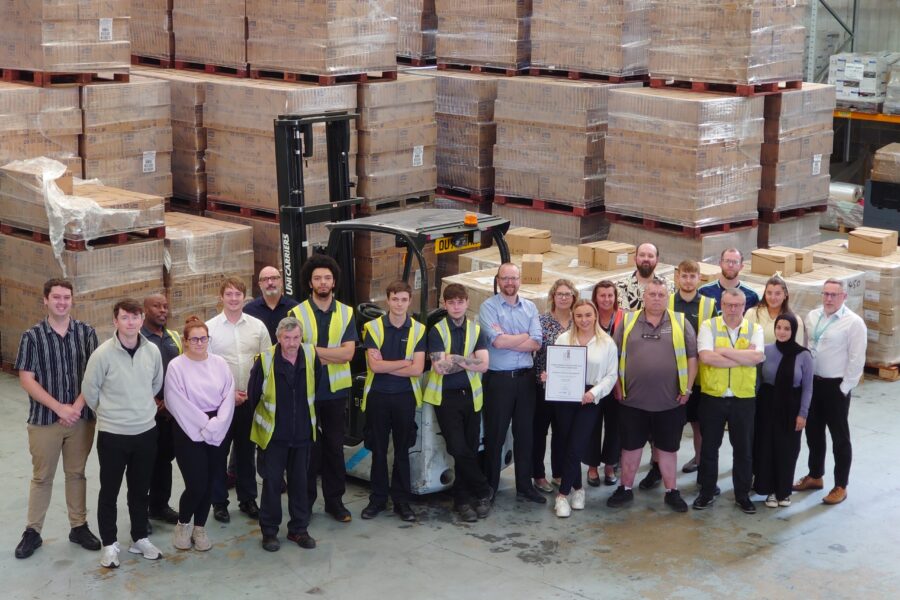 Members of the Procurement Services team at Leeds Teaching Hospitals, pictured in the distribution warehouse with stacks of pallets and boxes in the background. One of the staff members is holding a certificate of customer service excellence.