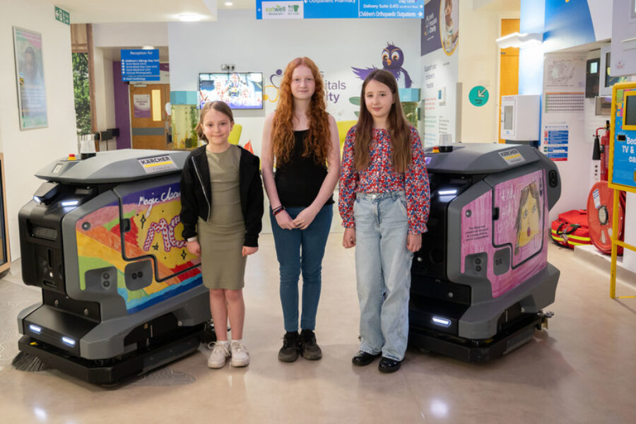 Photo of three children in the middle of two cleaning robots displaying the children's designs