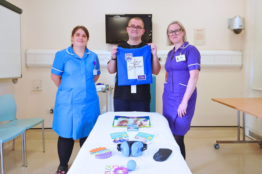 Two nurses in uniform and a young man with glasses stand around a patient bed in a clinical room. The man is holding a blue tote bag and on the bed are a number of sensory items and distraction aids.