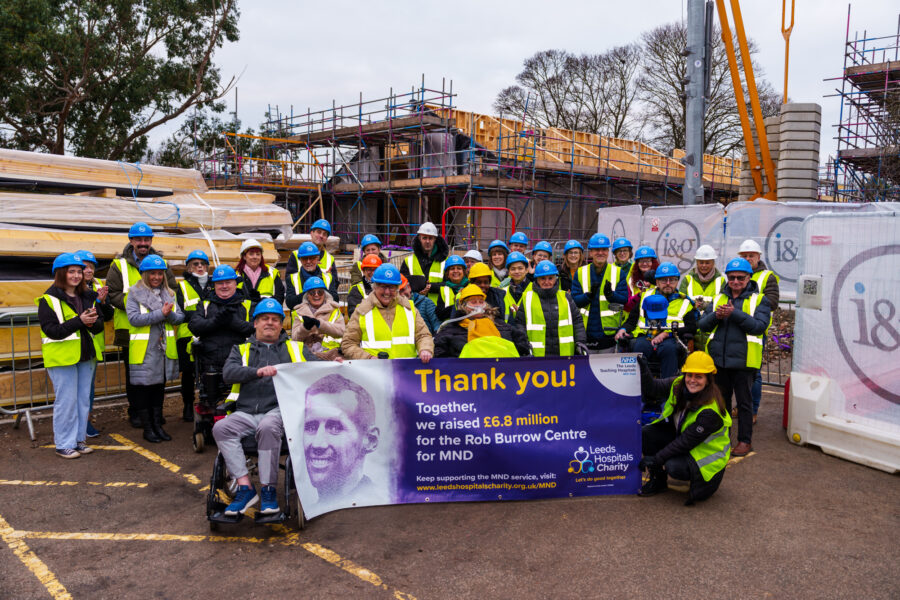 Group of people on a construction site wearing hard hats and high vis jackets, holding banner that says Thank you to all the supporters for raising the £6.8 million funds for the Rob Burrow Centre for Motor Neurone Disease