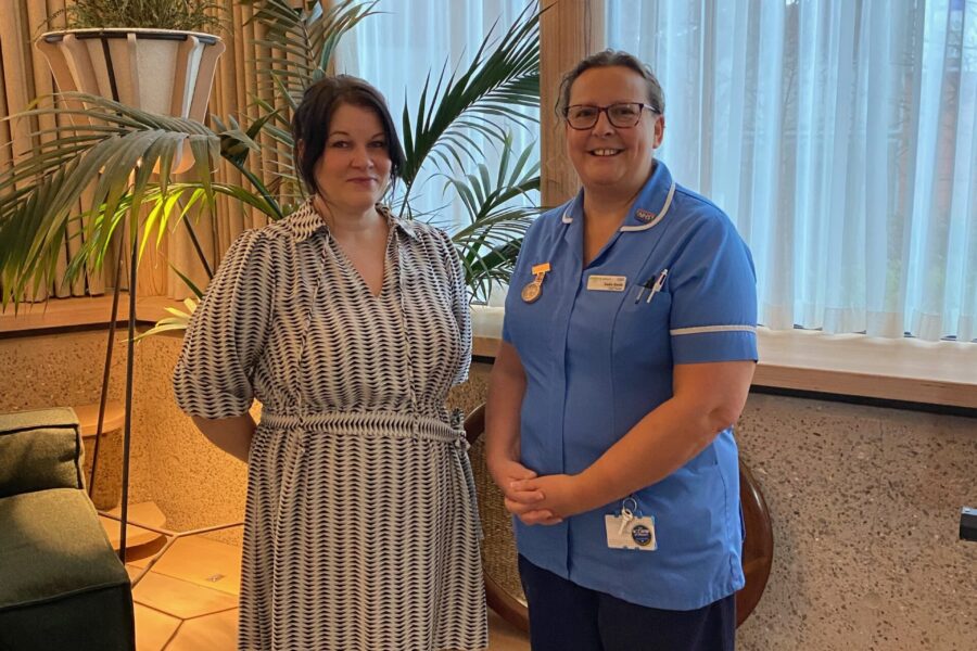 Two people standing in a dimly lit room in front of some plants. One is wearing a clinical uniform