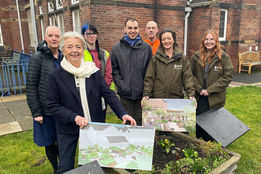 Group of people standing behind a planter tub in a garden in front of a brick building. Two people are holding signs showing pictures of a garden