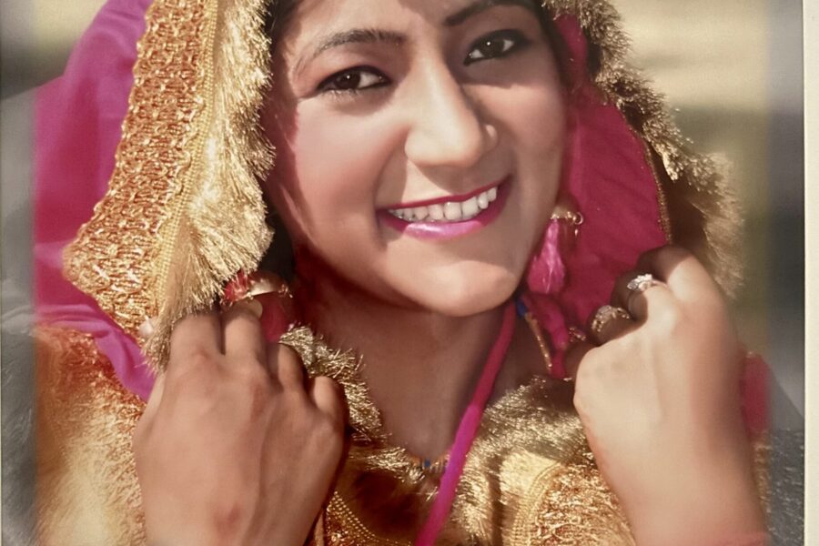 A smiling woman wearing traditional South Asian attire, including a bright pink and gold dupatta with intricate detailing, matching bangles, and a maang tikka (forehead ornament). She holds the sides of her dupatta near her face, radiating joy and confidence."