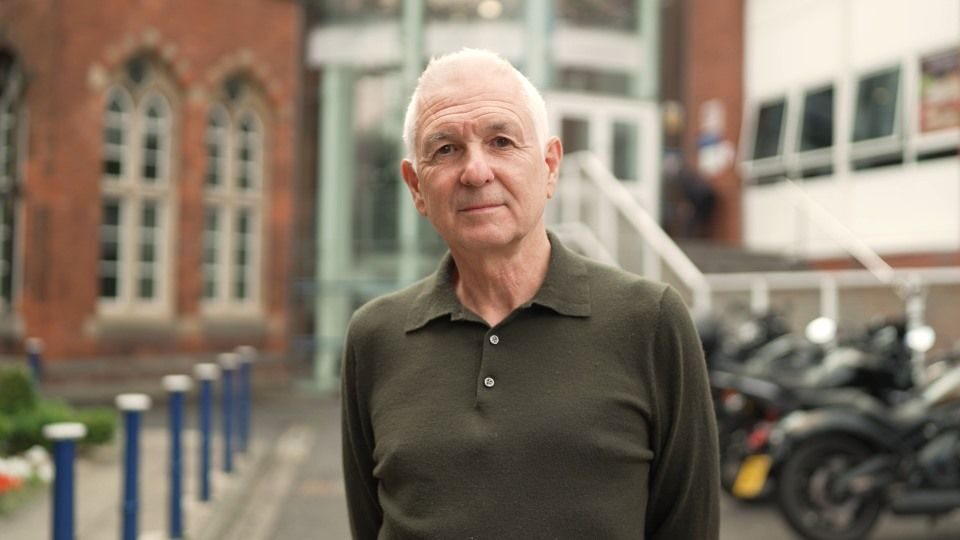 A man in a dark green shirt stood in front of a hospital building