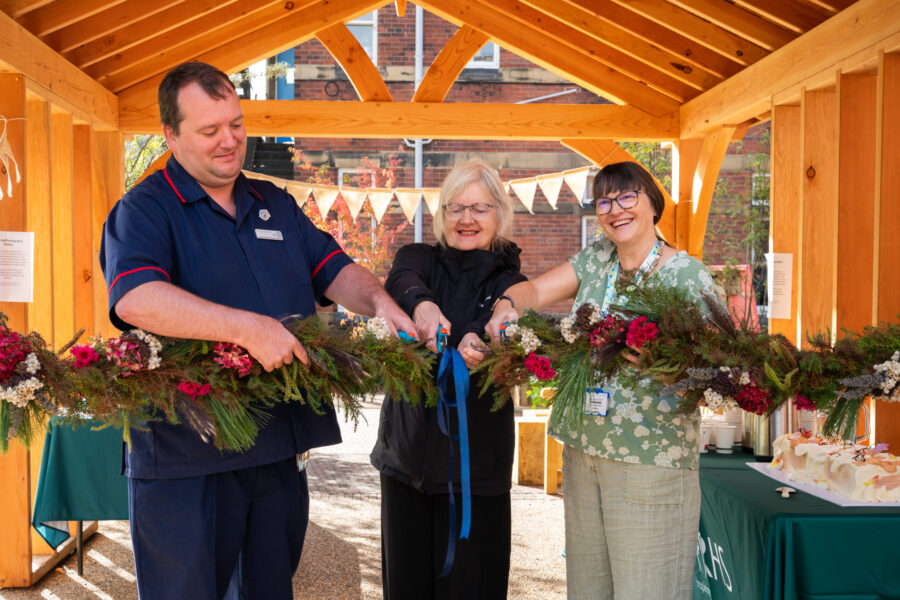 Three people standing in front of a wooden shelter and cutting a garland.