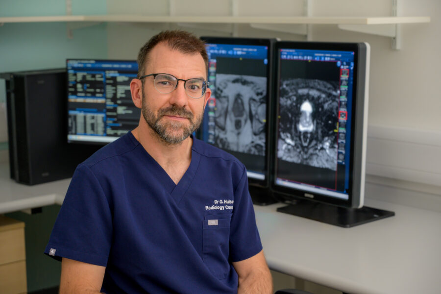 An image if Dr Oliver Hulson sitting facing the camera with medical scan images on computer monitors behind him.