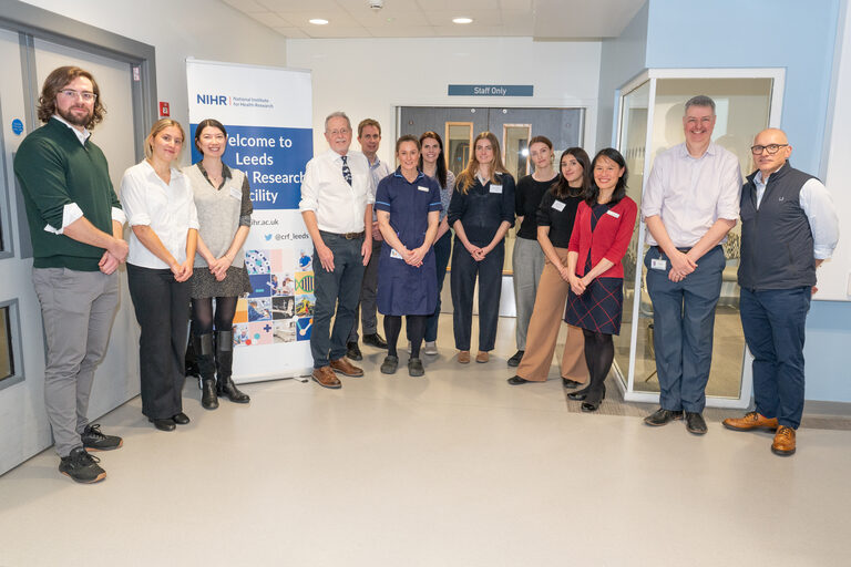 A photo of guests and delegates alongside members of the Trust's research team standing outside the NIHR Leeds Clinical Research Facility at St James, next to a banner for the facility.