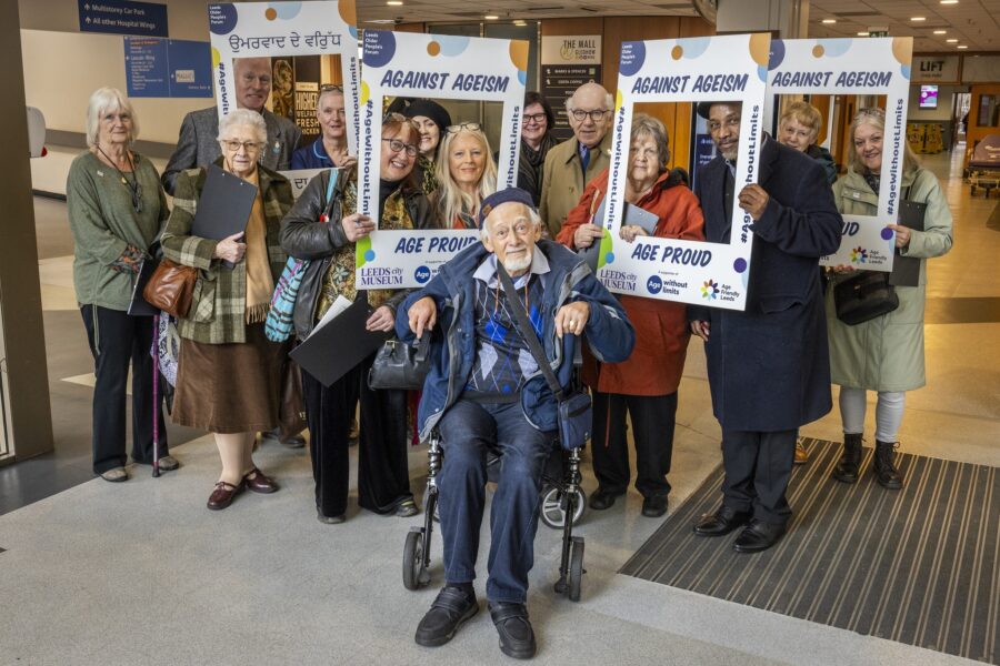 A group of people stood in a hospital lobby, some holding selfie frames branded Age Friendly Leeds. An older man in a wheelchair is at the front of the group.
